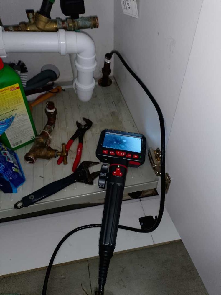 A handheld borescope camera with a lit screen, inspecting pipework in the cupboard beneath a sink