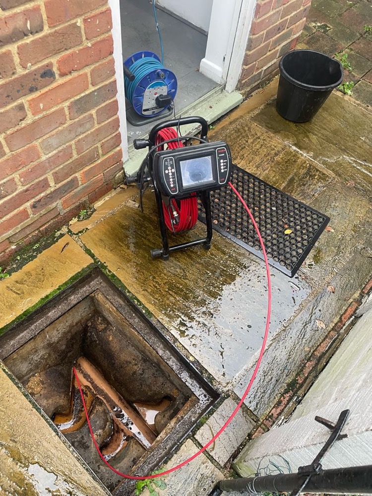 A drain camera reel with a screen, set up beside an open drain chamber with pipework visible inside