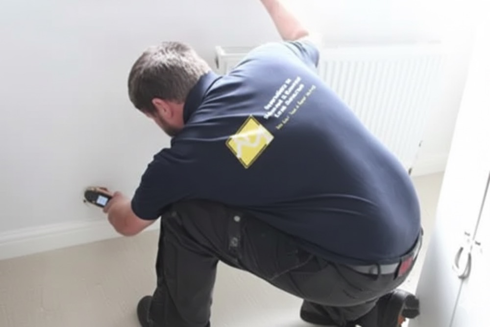 An ADI engineer kneeling at a skirting board, holding a handheld moisture meter against the wall beside a radiator