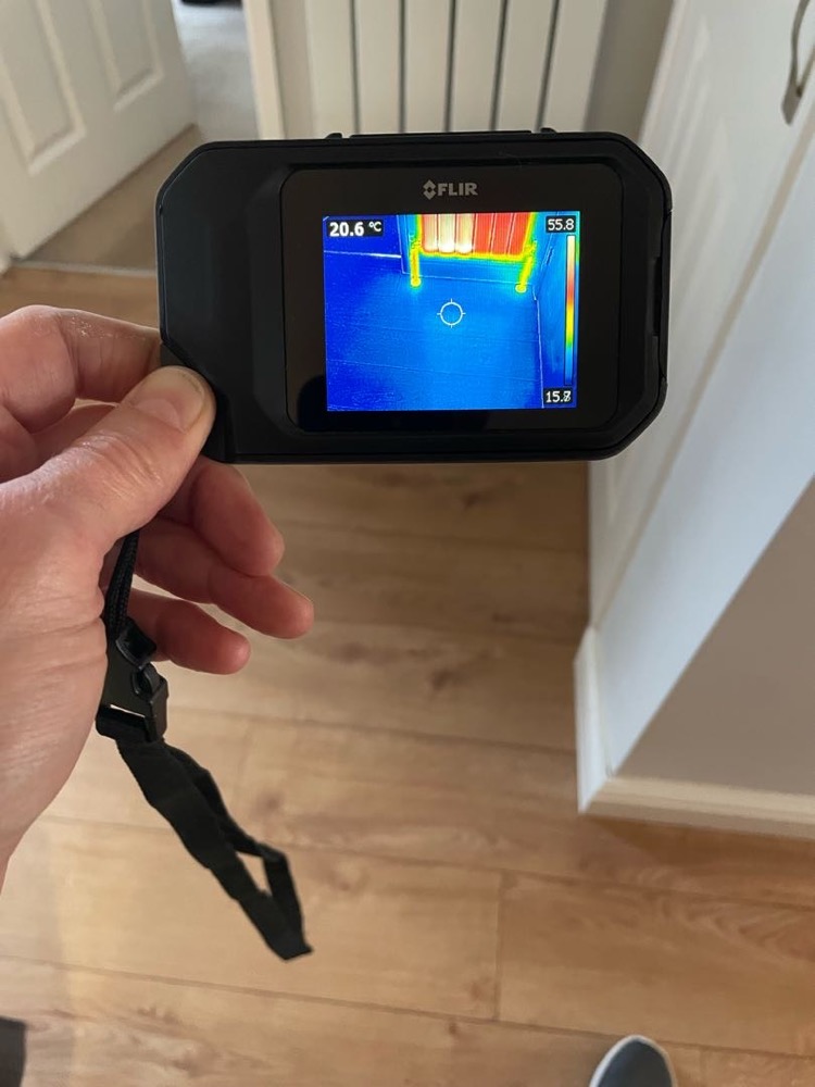 A handheld FLIR thermal camera showing hot pipework in red and yellow against a cool blue floor